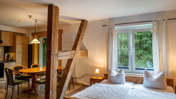 A bedroom with a wooden bed, a window with curtains, a dining area with a table and chairs, and a kitchen in the background.