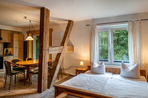 Un dormitorio con cama de madera, una ventana con cortinas, una zona de comedor con mesa y sillas, y una cocina al fondo.