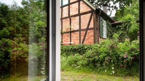 Blick durch ein Fenster auf ein traditionelles Fachwerkhäuschen mit Garten.