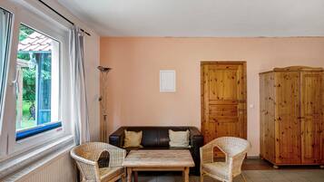 Una habitación con sofá, dos sillones de mimbre, una mesa de madera, radiador, ventana con cortinas y armario de madera.