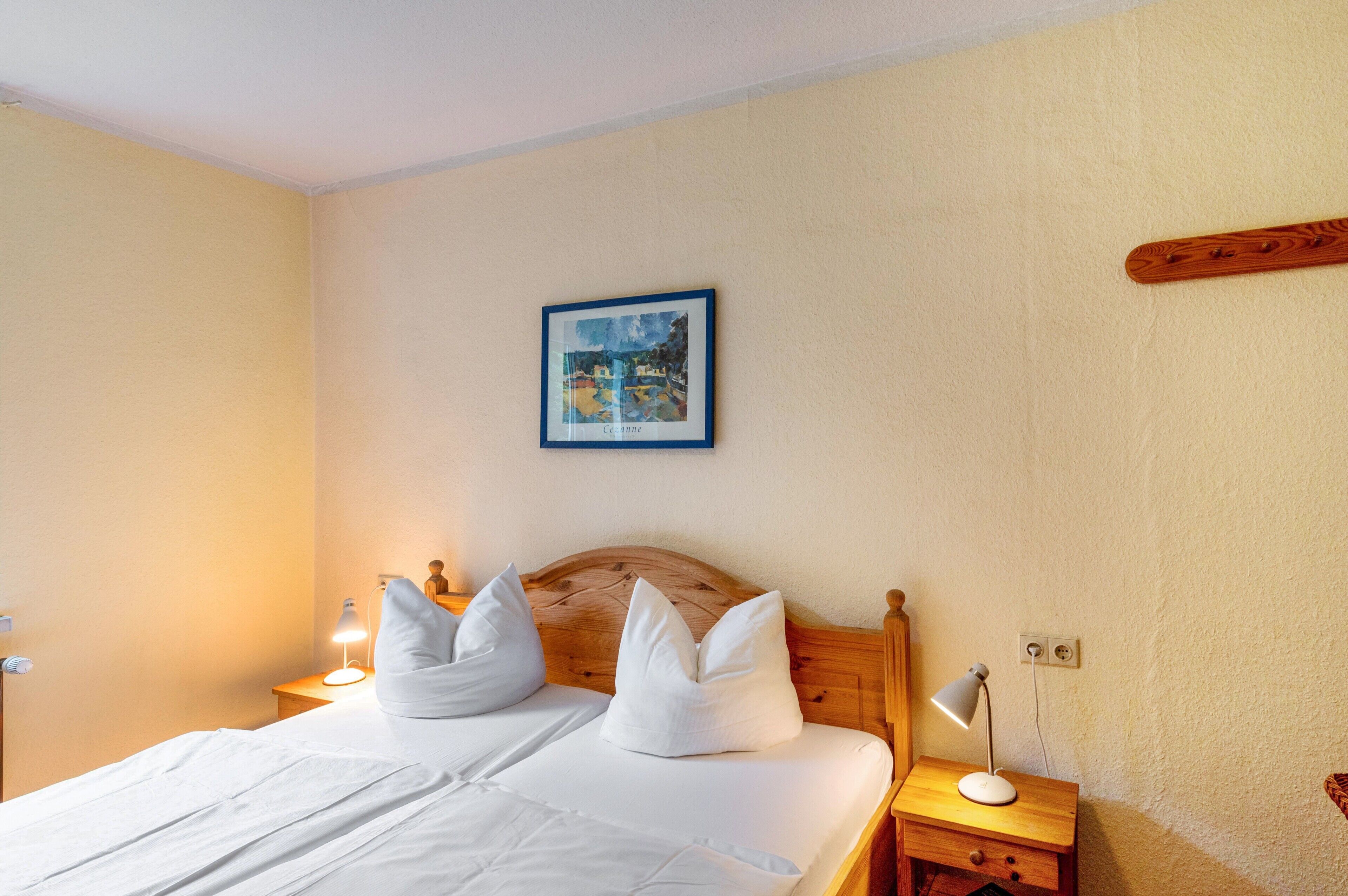 Una cama bien hecha con sábanas y almohadas blancas, un cabecero de madera, una lámpara de noche, un cuadro en la pared y una mesita de noche de madera con una lámpara.