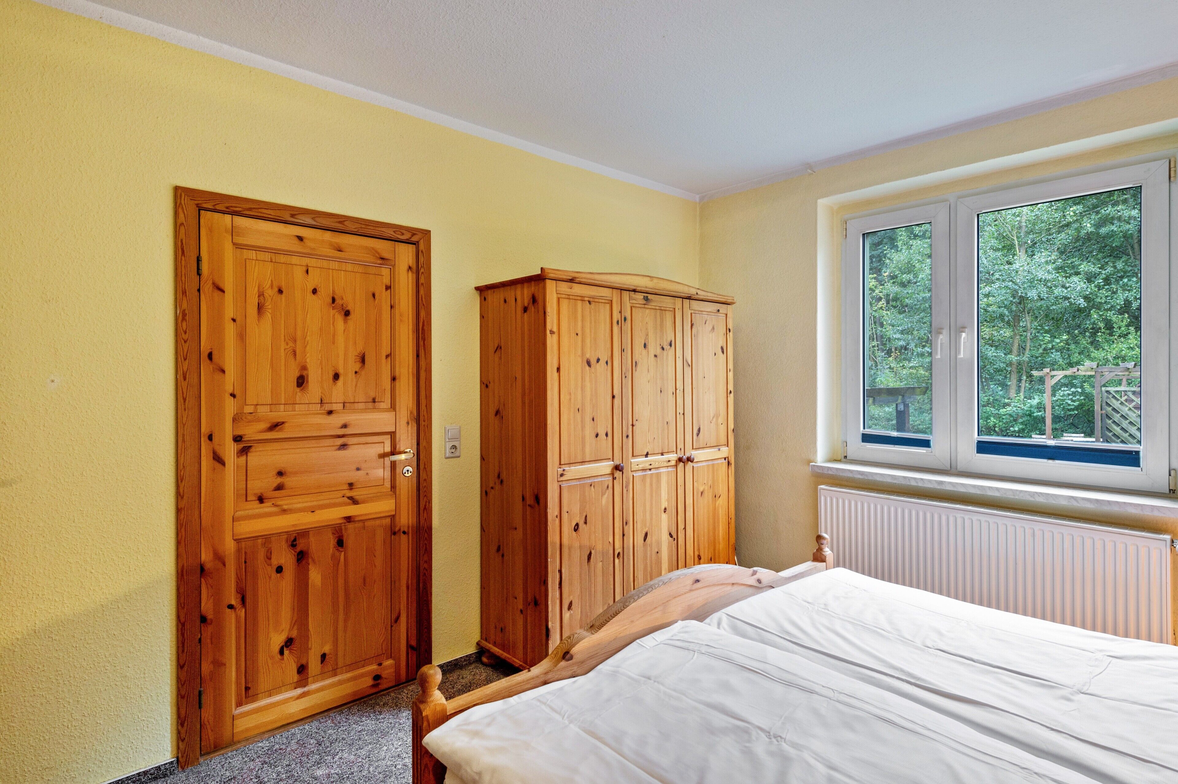 A bedroom with a bed, wooden wardrobe, and a window with a view of greenery.