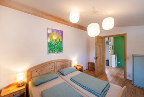 A bedroom with a wooden headboard, two beds with blue bedding, a nightstand with a lamp, a painting on the wall, and a green door leading to another room.