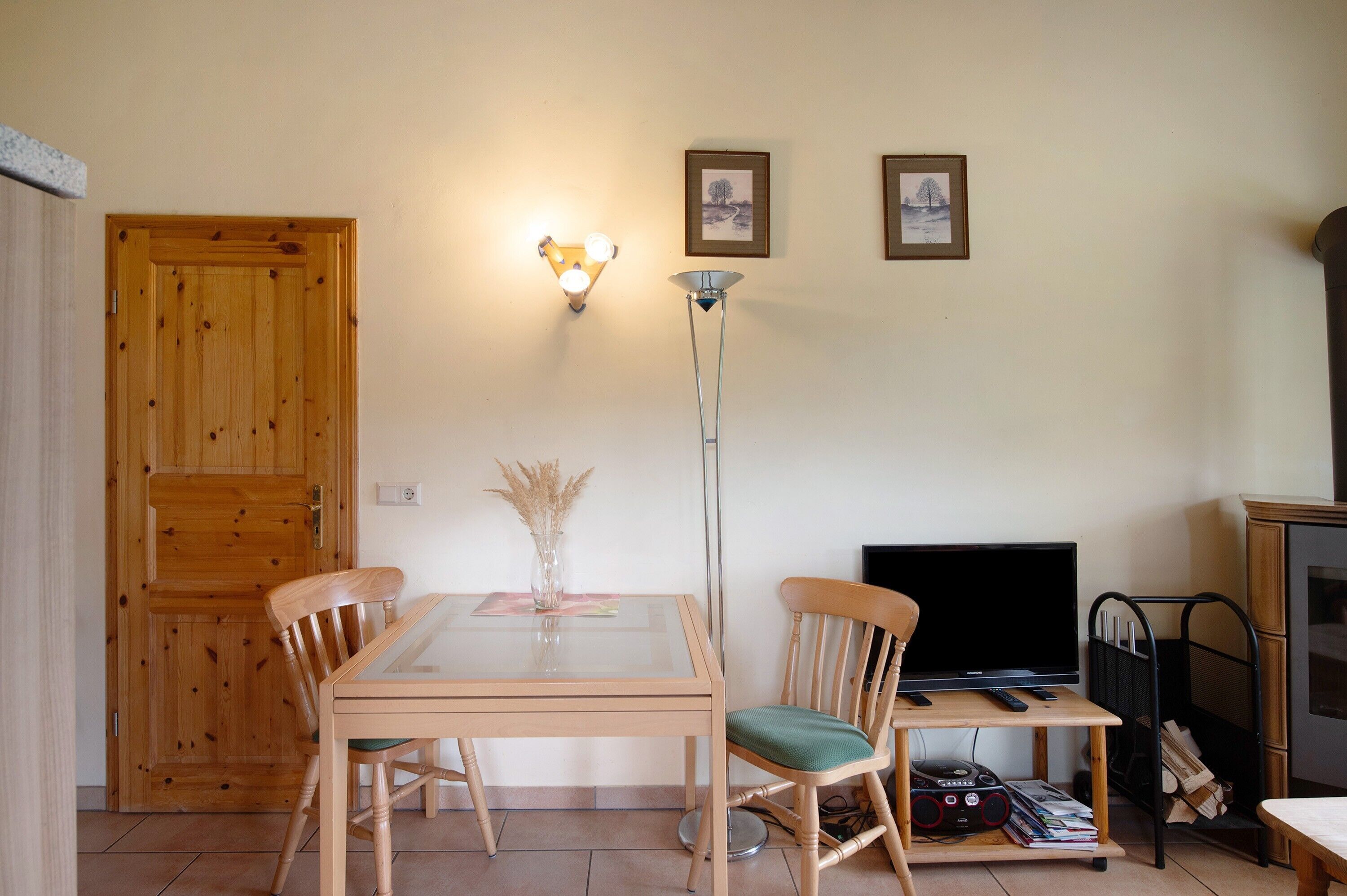 Un área de comedor con una mesa y sillas de madera, un jarrón con plantas secas, una lámpara de pie y un televisor sobre un mueble.