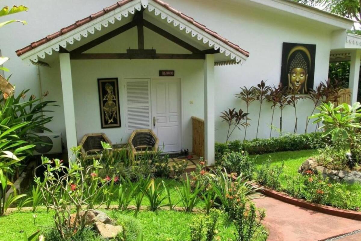 A white building with a tiled roof, a porch with two wicker chairs, and a Buddha statue on the wall.