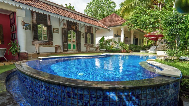 Une piscine aux carreaux de mosaïque bleus, entourée d’un jardin où poussent différentes espèces de plantes, et une maison avec un toit recouvert de tuiles.