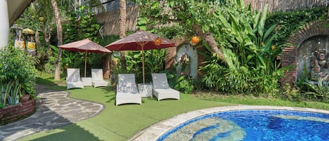 A swimming pool with a tiled edge, surrounded by artificial grass and a brick wall. There are two white lounge chairs and two umbrellas.