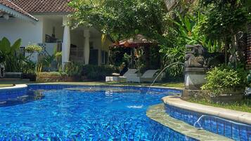 A swimming pool with clear blue water, surrounded by lush greenery and a building with a tiled roof.