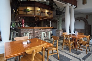 A restaurant interior with wooden tables and chairs, a bar area with stools, and a brick wall.