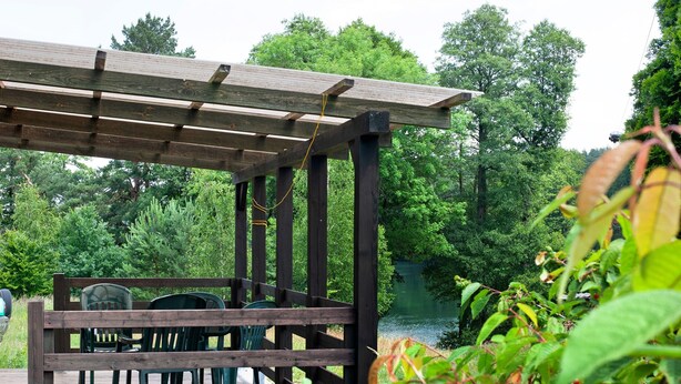 Una pérgola de madera con sillas y una mesa, con vistas a un lago rodeado de árboles.