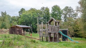 A wooden play structure with a slide and climbing elements, surrounded by trees and grass.