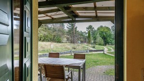 An outdoor patio with a table and chairs, a covered area with a roof, and a view of a grassy area with trees.