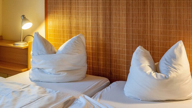 A hotel room with two beds, each with a pillow, a bedside lamp, and a wooden headboard.