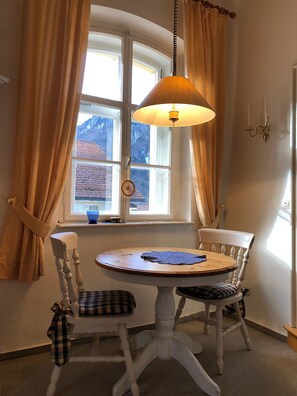 Quarto com mesa redonda, duas cadeiras, luminária pendente, janela com cortinas e luminária fixada na parede.