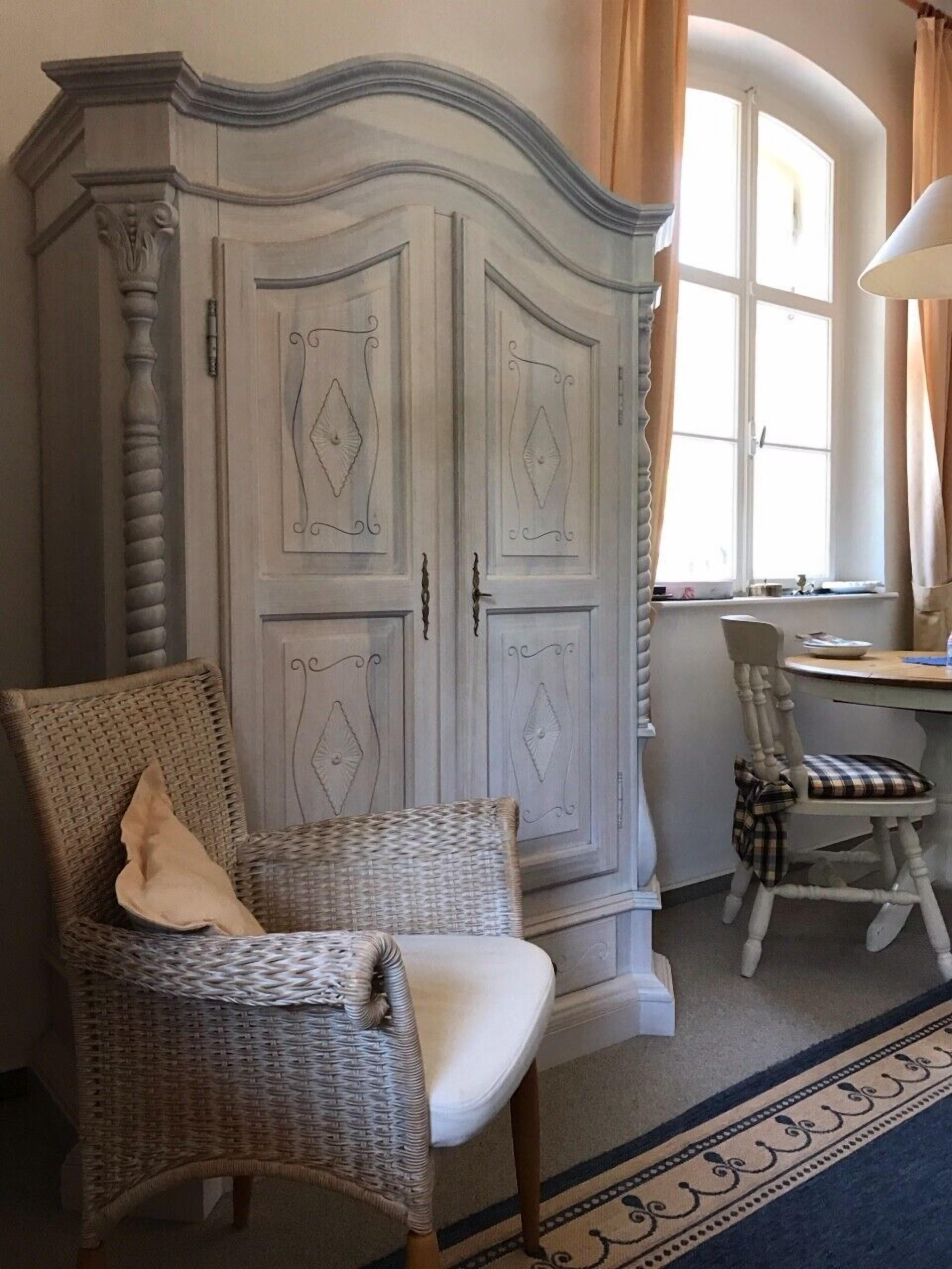 Een kamer met een grote witte kast, rieten stoelen, een klein tafeltje en een raam met gordijnen.