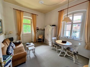 Quarto com um sofá marrom, uma mesinha, uma cadeira branca, um armário e duas janelas com cortinas.