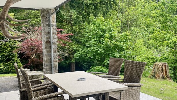 An outdoor patio with a table and wicker chairs, surrounded by greenery and a stone pillar.
