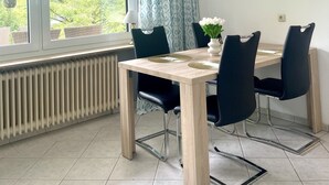 A dining area with a table, chairs, a radiator, a window with curtains, and wall decorations.