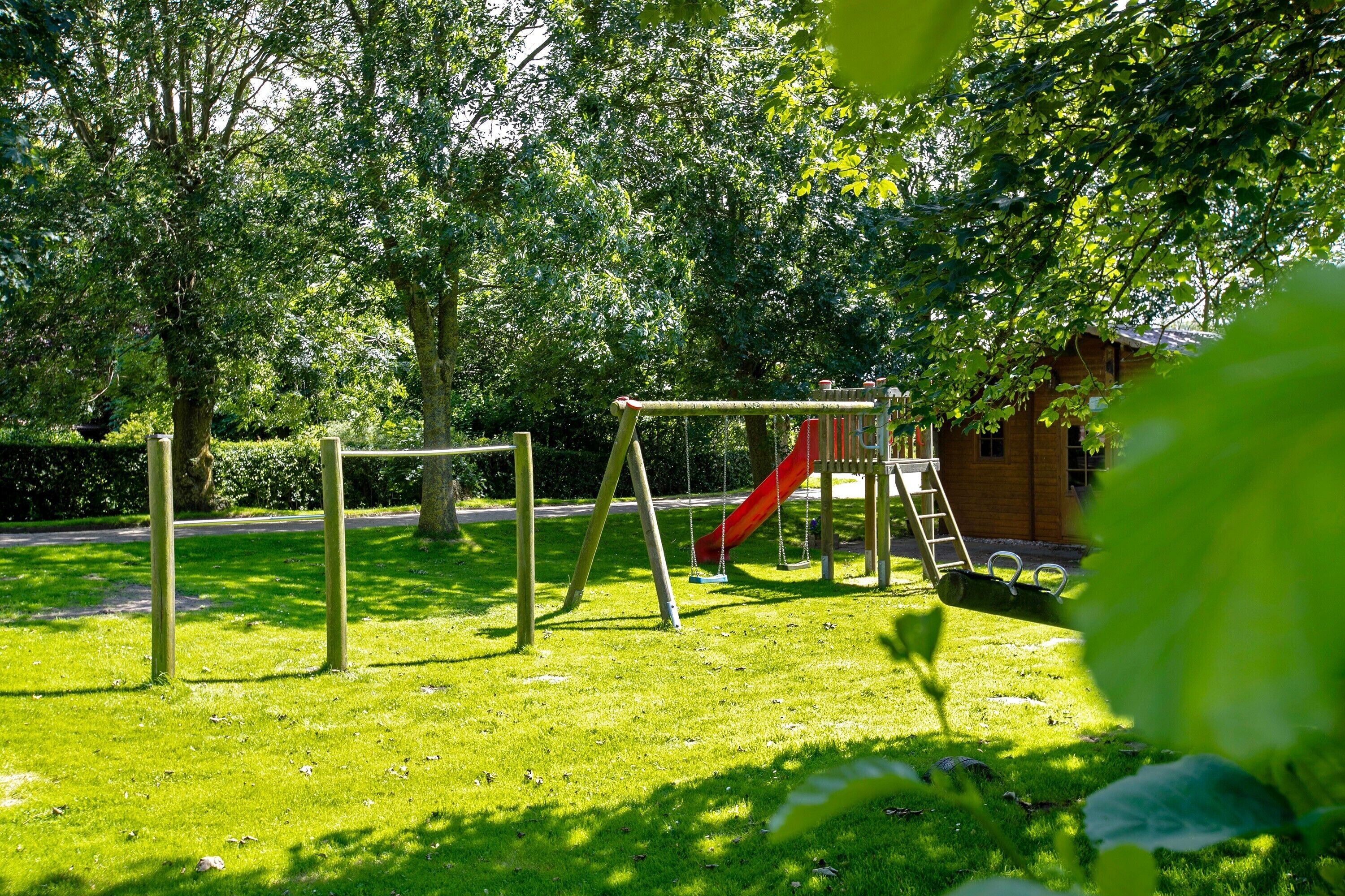 Ein Spielplatz mit Schaukeln und einer Rutsche, umgeben von Bäumen und einem Gebäude im Hintergrund.