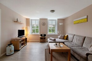 Un salon avec un canapé, une table basse, un téléviseur posé sur un meuble, une lampe et une horloge au mur.