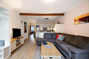 Uma sala de estar moderna com sofá, mesa de centro, televisão e cozinha ao fundo.