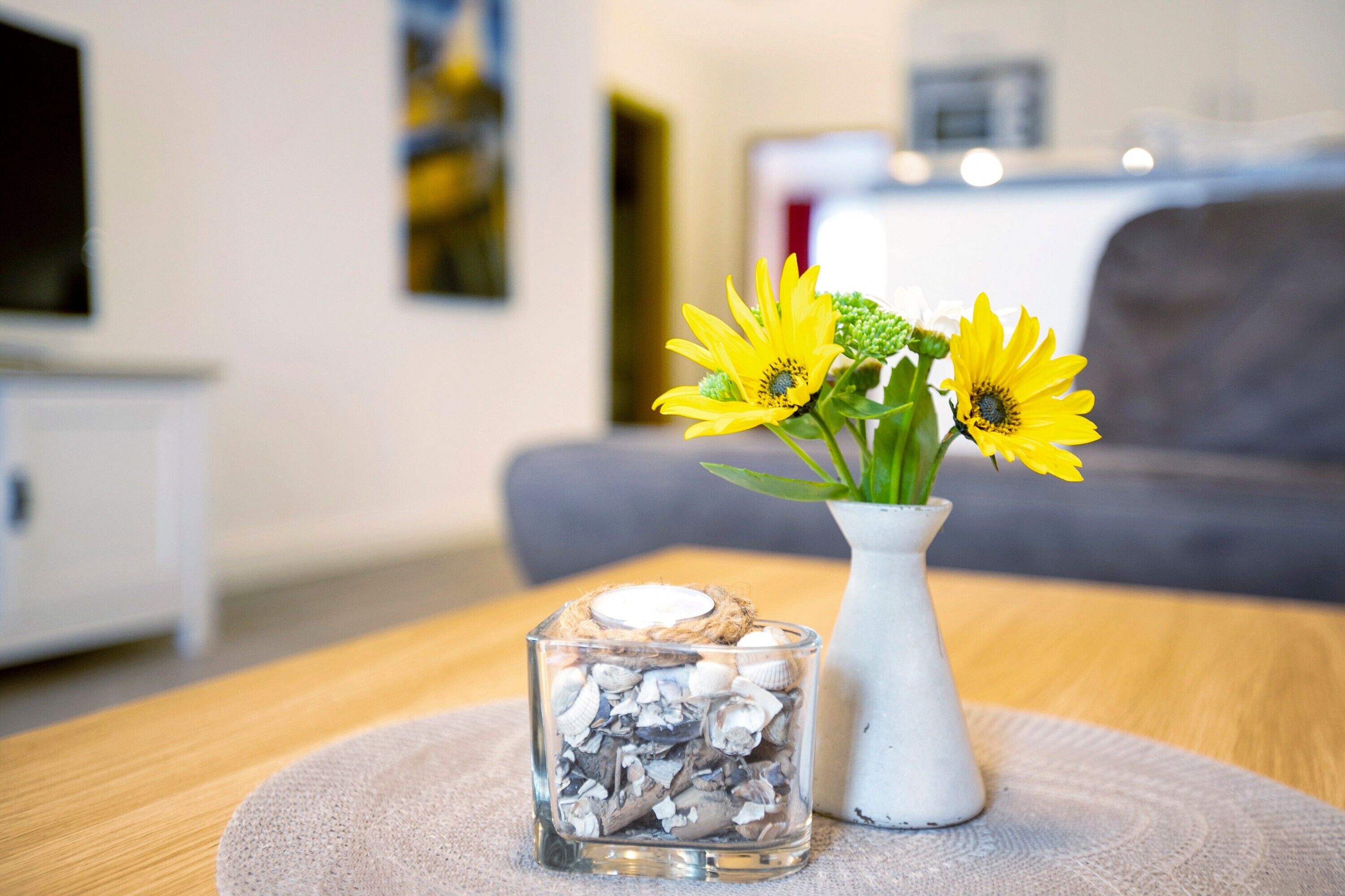Uma sala de estar com uma mesa de centro de madeira, um vaso branco com flores amarelas e um recipiente de vidro com conchas.
