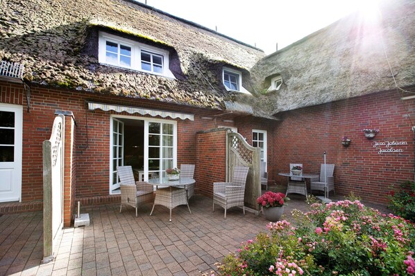 A brick building with a thatched roof, a patio with wicker furniture, and a garden with pink flowers.