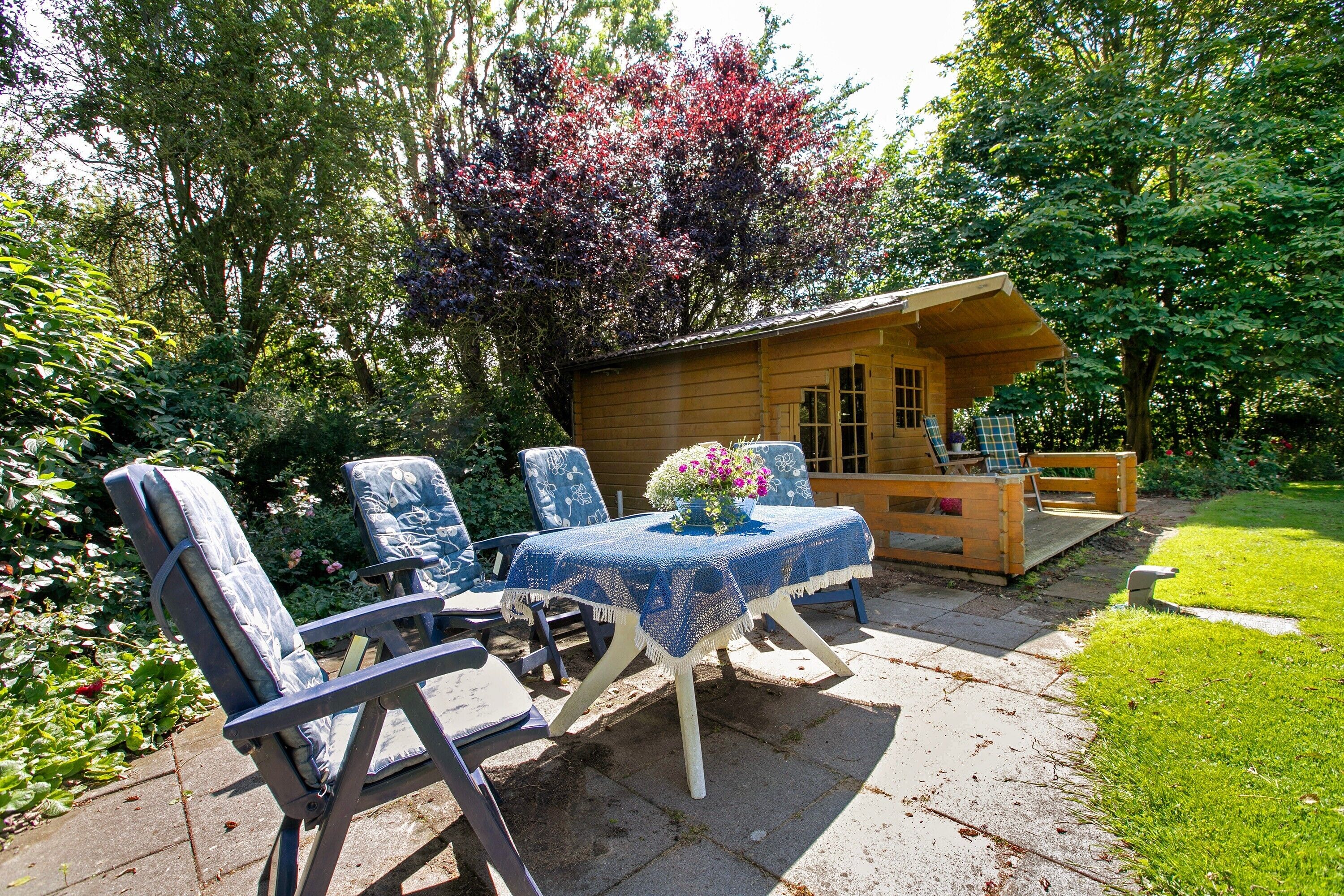 Une cabane en bois avec une véranda, une table entourée de chaises et un jardin agrémenté de diverses plantes et arbres.