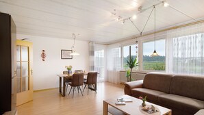 Un vaste espace de vie comprenant un canapé, une table à manger, des chaises et une grande fenêtre donnant sur un paysage verdoyant.