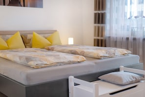 Una cama doble con colchón gris y cabecera blanca, una mesita de noche con lámpara blanca, y una ventana con cortinas traslúcidas.