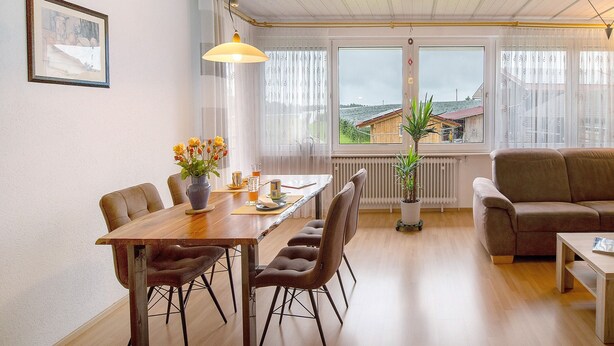 Un coin repas avec une table en bois, des chaises et un vase de fleurs. On y trouve également un canapé et une fenêtre donnant sur des immeubles.