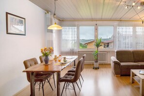 Un comedor con una mesa de madera, sillas y un florero. Hay un sofá y una ventana con vistas a edificios.