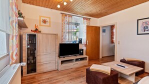En moderne stue med træloft, fjernsyn på træstel, et glaskabinet og et lille bord med en bog.