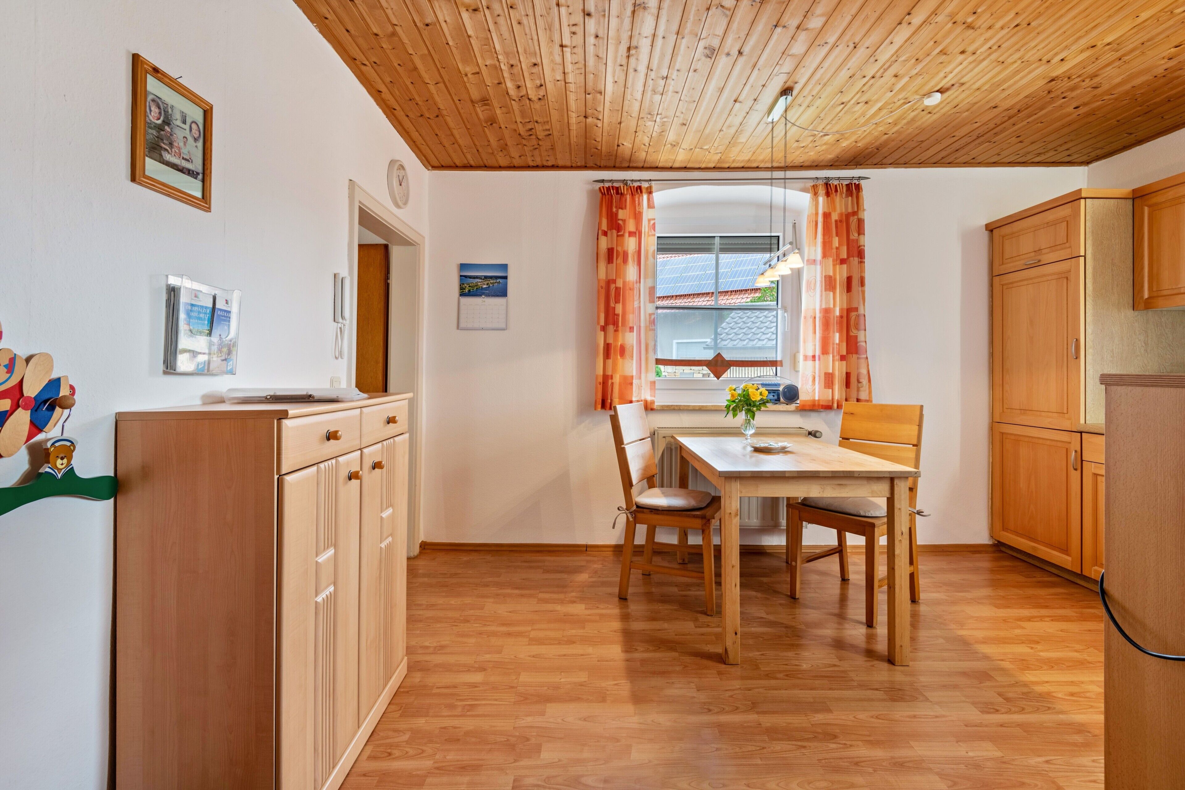 Una cocina con armarios de madera, una mesa de comedor con sillas, un ventanal con cortinas y un reloj en la pared.