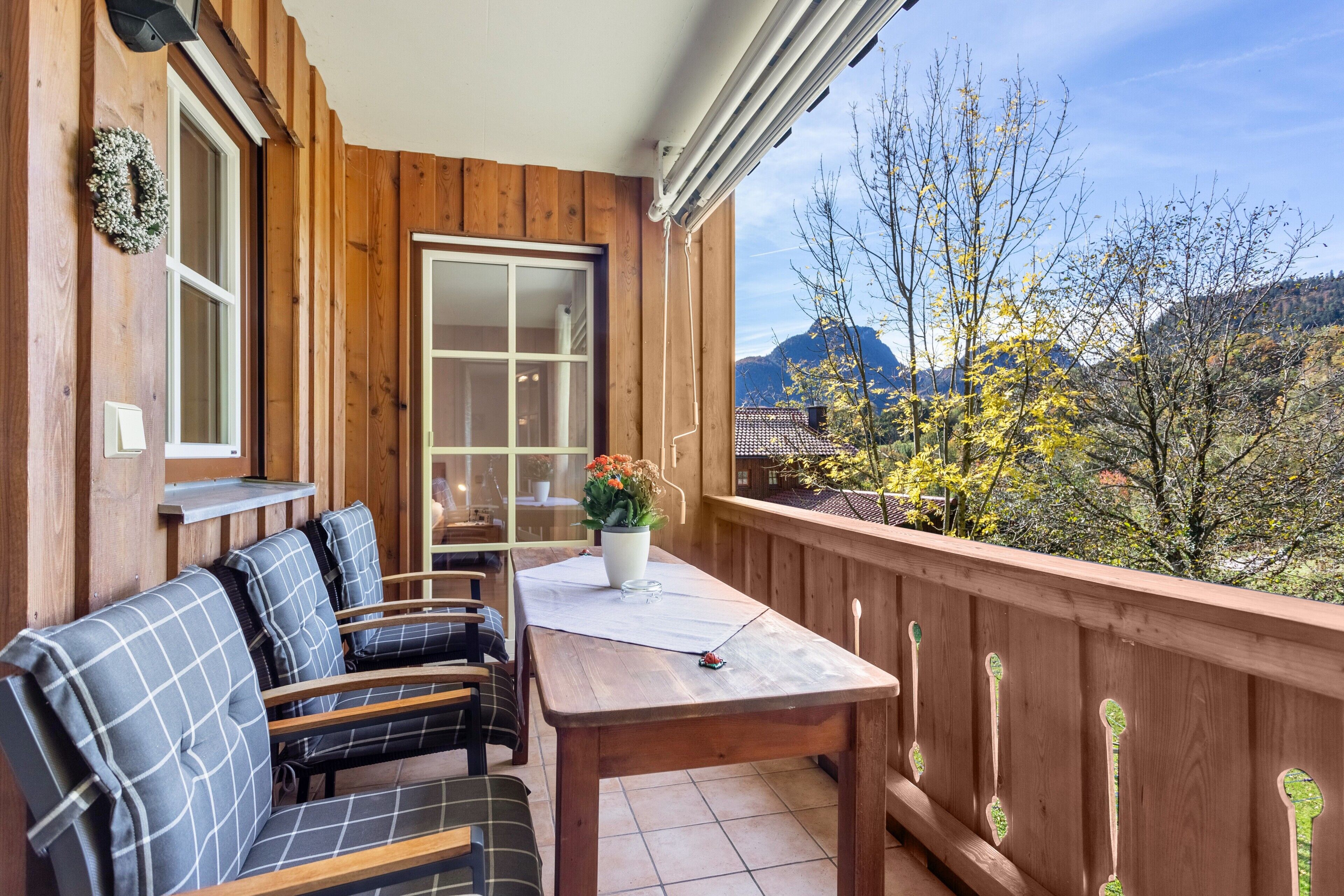 A wooden balcony with a table and chairs, a window, and a view of trees and mountains.