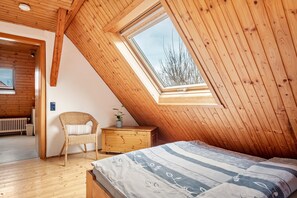 Une pièce avec un plafond en pente, des murs en bois, un lit avec une literie à motifs, une chaise en osier, une petite table de chevet en bois et une fenêtre donnant sur les arbres.