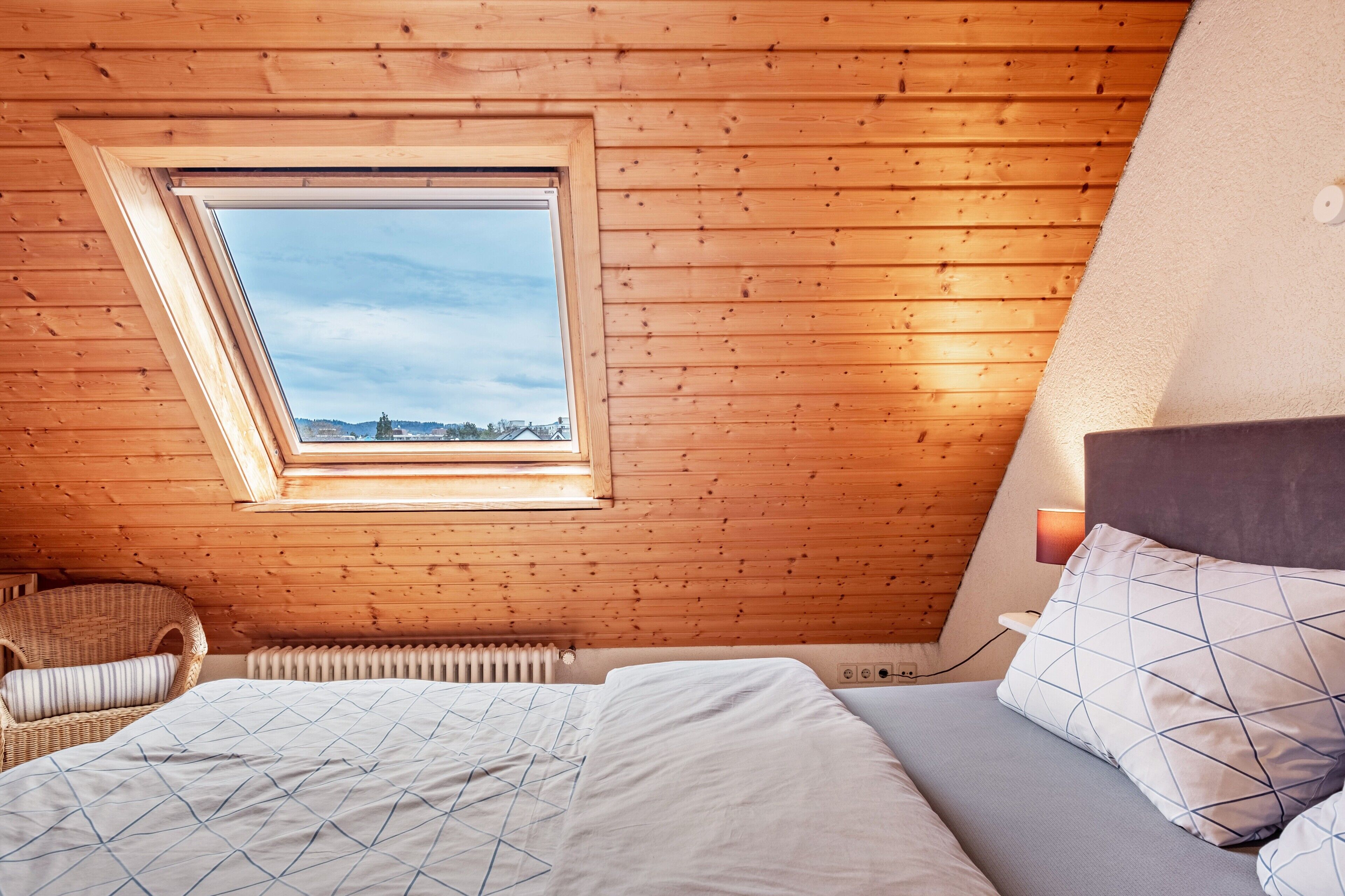 Une chambre avec un plafond en pente, un puits de lumière, un lit avec un coussin à motifs, une chaise et un radiateur.