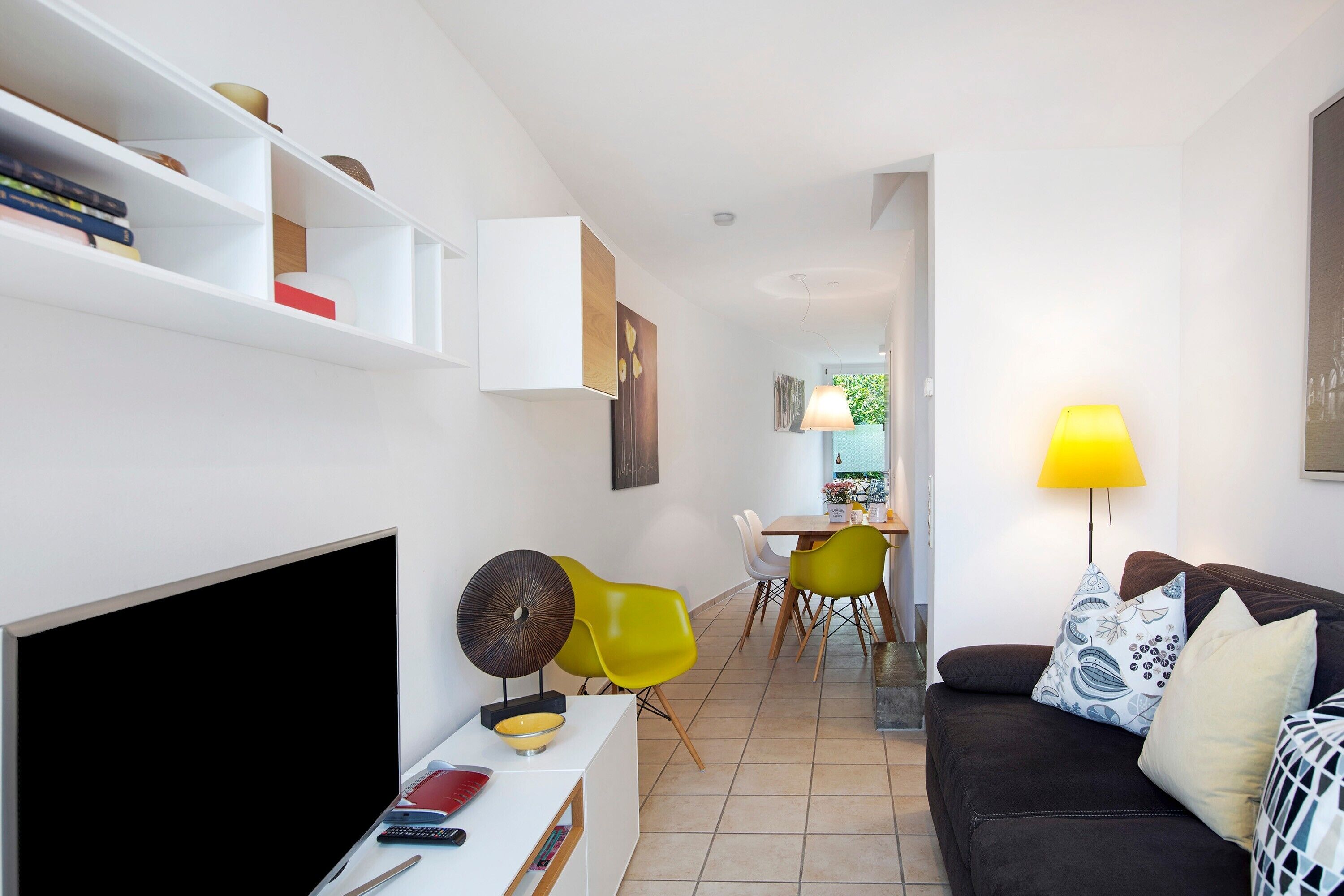 Una sala de estar moderna con un televisor de pantalla plana, un comedor con sillas amarillas y un sofá con cojines estampados.