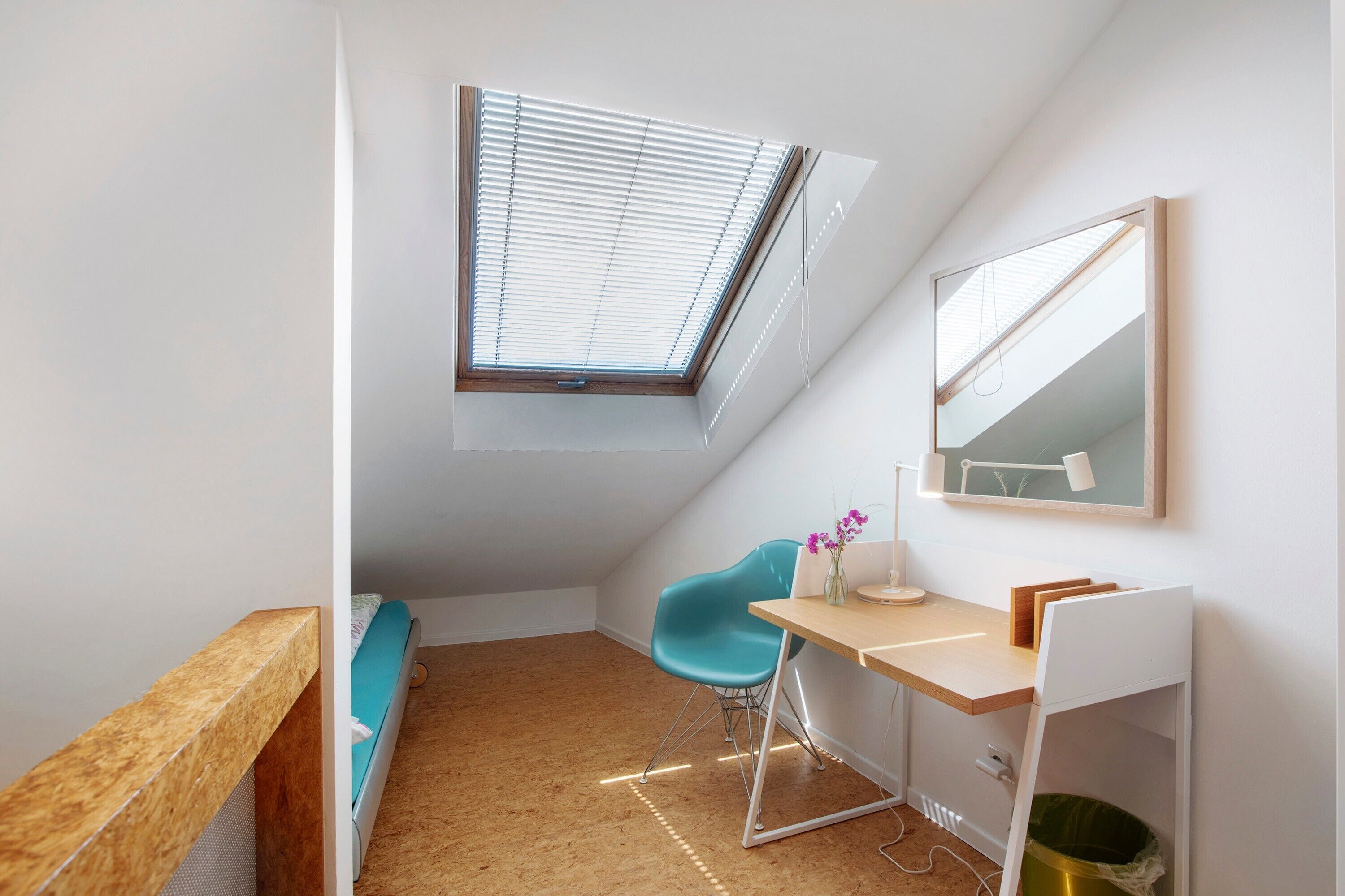 Een kleine, moderne kamer met een schuine kap, dakraam, bureau met stoel, bed en spiegel aan de muur.