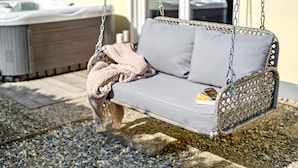 A wicker swing chair with cushions, a blanket, and a bottle on a gravel surface.