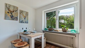 A dining area with a table set for two, a window with a view of greenery, and two paintings on the wall.
