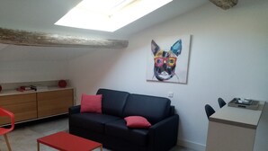 Un espace de vie compact avec un canapé noir, des coussins rouges, une table basse en bois, une chaise rouge, un petit bureau et un tableau représentant le visage d’un chat accroché au mur.