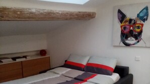 Une chambre avec une lucarne, un lit recouvert d’une couvre-lit grise et rouge, une tête de lit noire et un tableau représentant un chien accroché au mur.