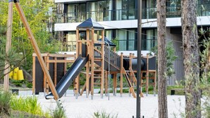 Un terrain de jeux avec un toboggan et des structures d’escalade, entouré d’arbres et avec un bâtiment moderne en arrière-plan.