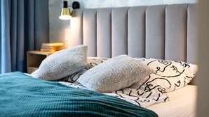Un lit bien fait, recouvert d’une couverture turquoise, avec deux oreillers moelleux et un coussin à motifs. Une lampe de chevet est allumée, et on aperçoit un mur orné d’un motif discret.