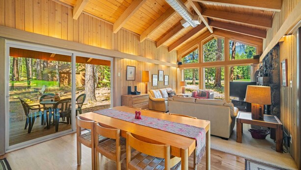 Una cabaña de madera espaciosa con comedor, sala de estar con chimenea y ventanas grandes que ofrecen vistas del bosque circundante.