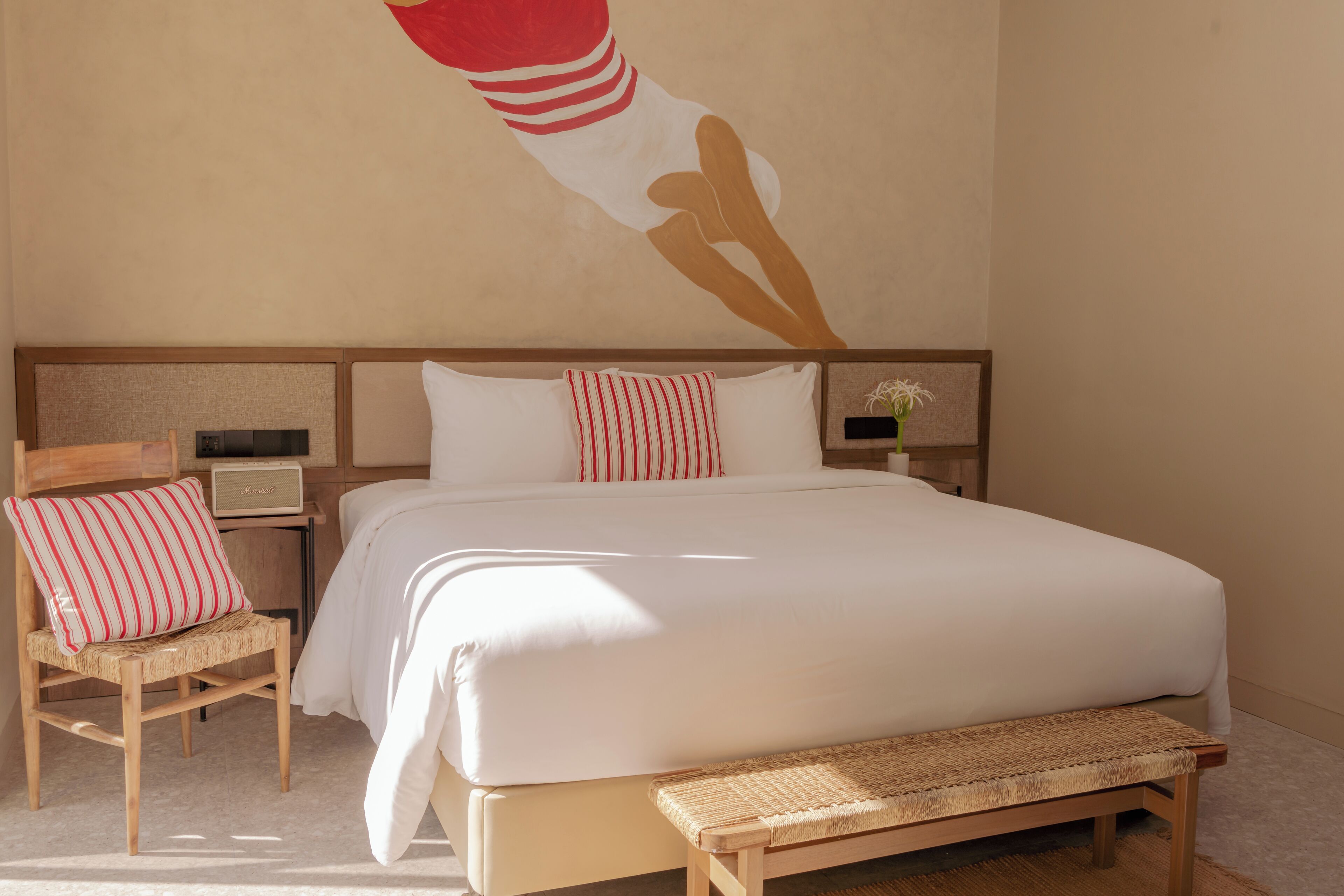 A neatly arranged bedroom with a large bed, a bedside table with a radio, a chair with a striped pillow, and a wall-mounted artwork.