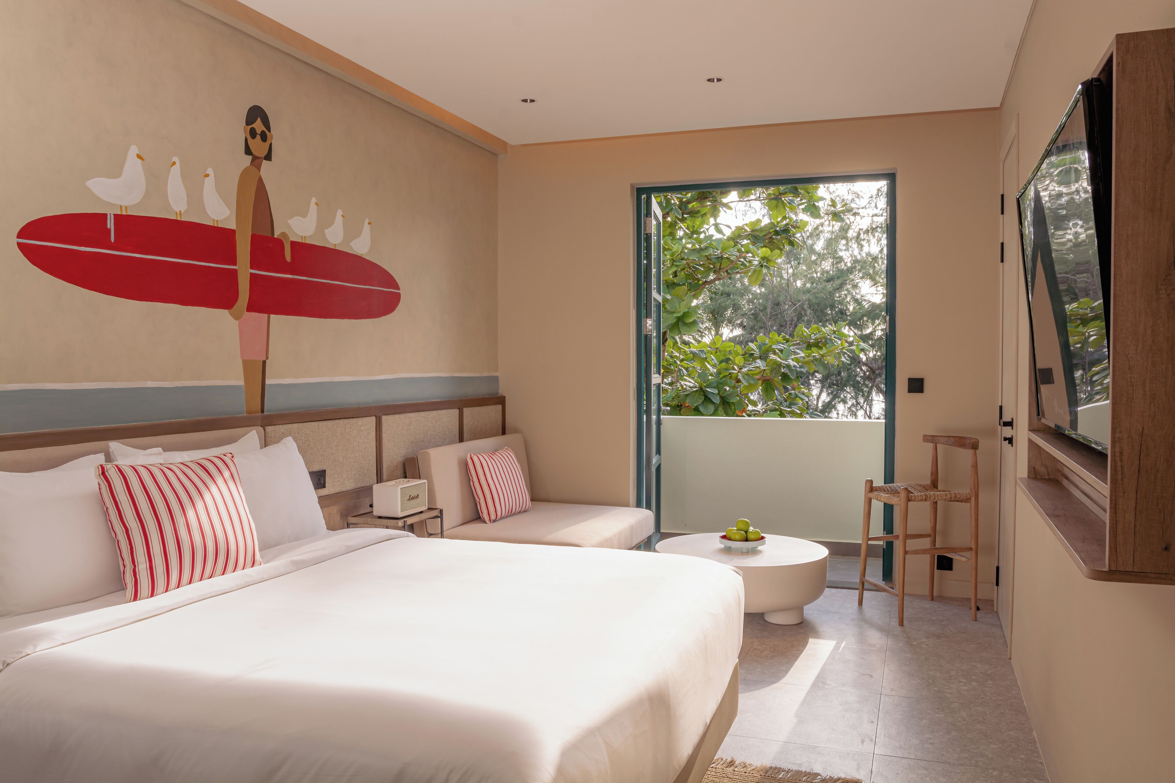 A hotel room with a large bed, a small sofa, a round coffee table, and a view of greenery through a large window.