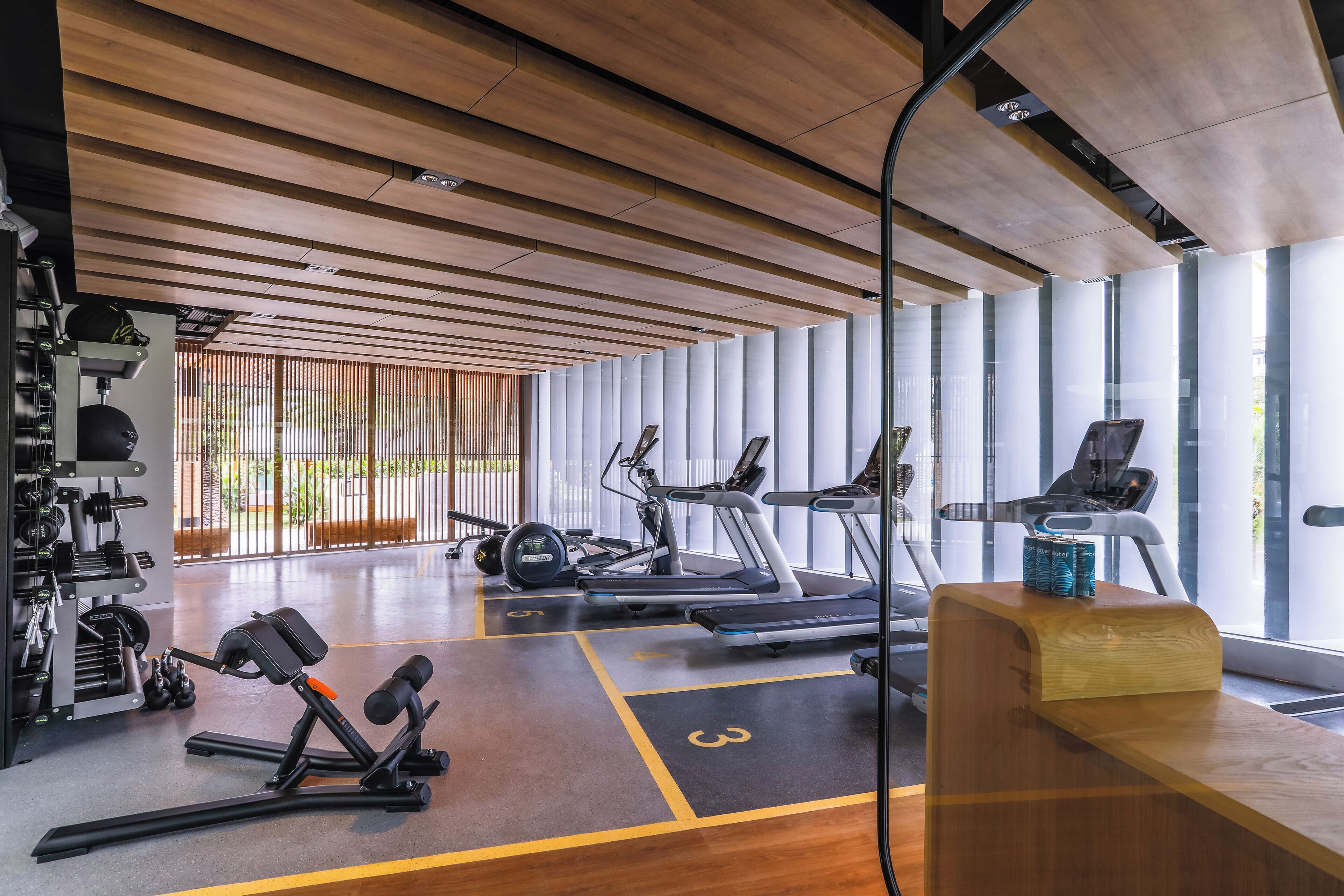 Un gimnasio moderno con equipamiento de ejercicio, una recepción de madera y ventanas amplias.