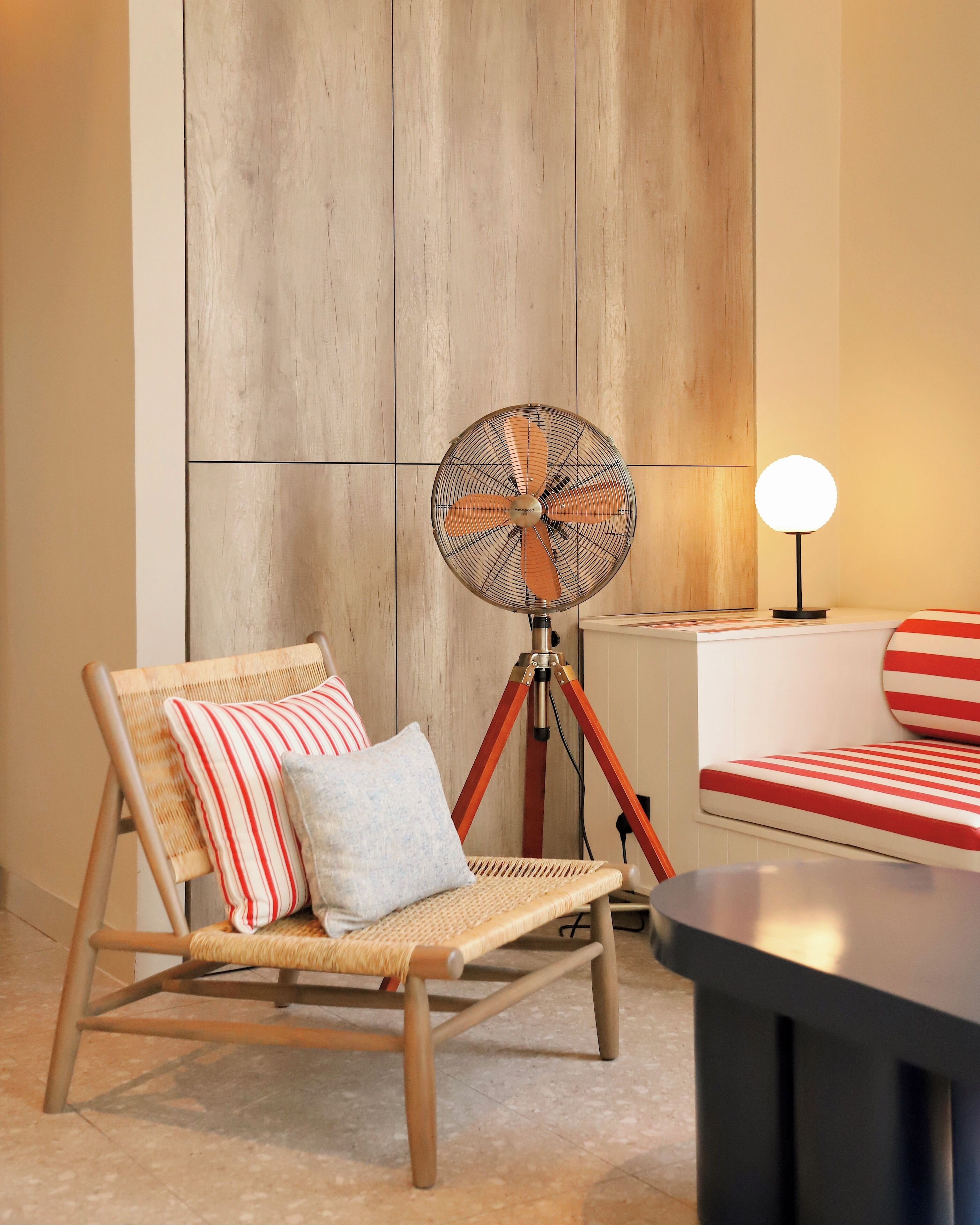 A room with a wicker chair, a tripod fan, and a striped cushion.